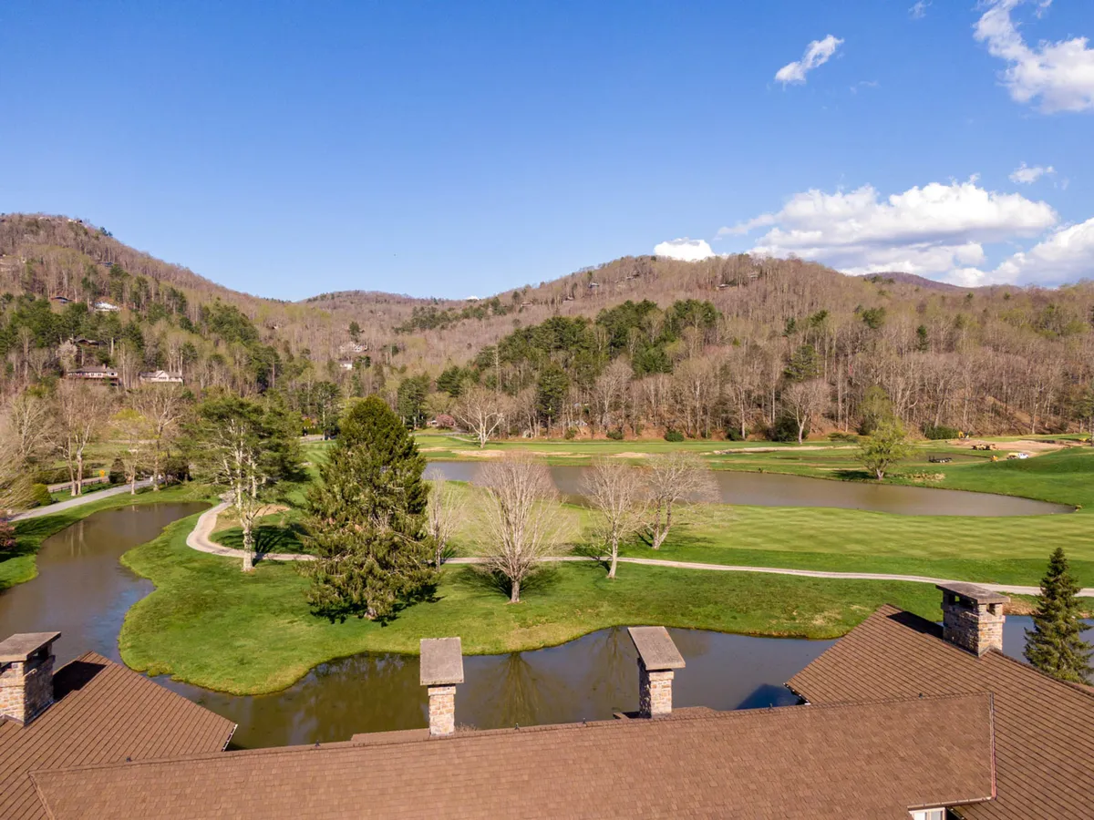A sweeping view captures a serene pond with a manicured golf course and dramatic mountain backdrop. The landscape is framed by a tree line, enhancing the sense of privacy and natural beauty. The setting is bathed in gentle light, suggesting either early morning or evening.