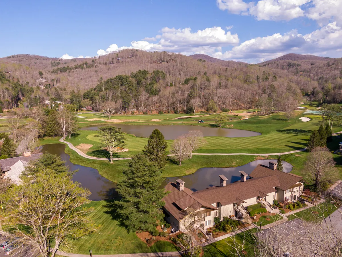 An expansive view of the condo community nestled among lush green fairways, tranquil ponds, and rolling mountains, all illuminated by the warm glow of a sunset. The golf course is the centerpiece, bordered by serene water features and dotted with walking paths, while elegant residential buildings are visible in the distance.