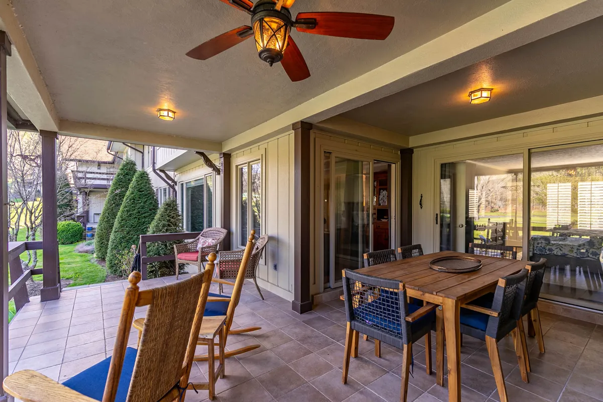 A cozy covered patio featuring a dining table and rocking chairs, with a ceiling fan overhead. The lush greenery surrounding the patio is visible through the open sides, creating a seamless connection to nature. The space is inviting for both quiet mornings and small gatherings, with ample seating and a relaxing ambiance.