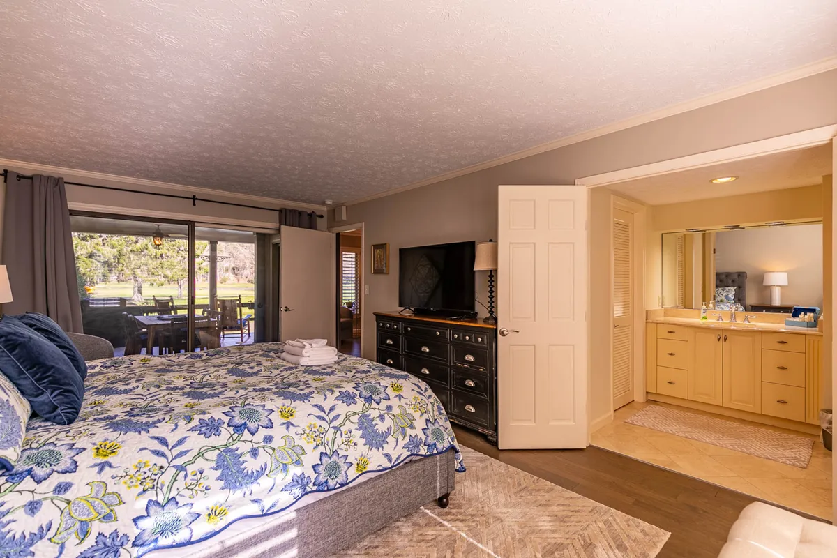 This image features a serene bedroom suite with a plush king bed adorned in floral bedding, a large TV, a vanity area, and a door that opens directly onto the patio. Natural sunlight pours in, and there's a sense of spaciousness and tranquility ideal for relaxation. The connection to the outdoors is subtly visible through the patio door.