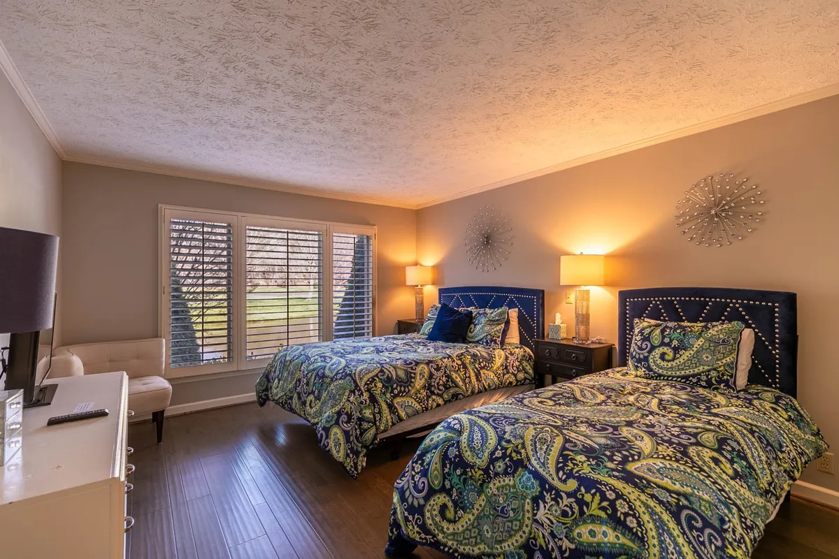 The second bedroom is shown with two beds adorned with colorful bedding, nightstands with lamps for each bed, a large window with shutters letting in natural light, and a cozy seating area. The setup feels inviting for families or friends sharing the space, and the décor is both calming and cheerful.