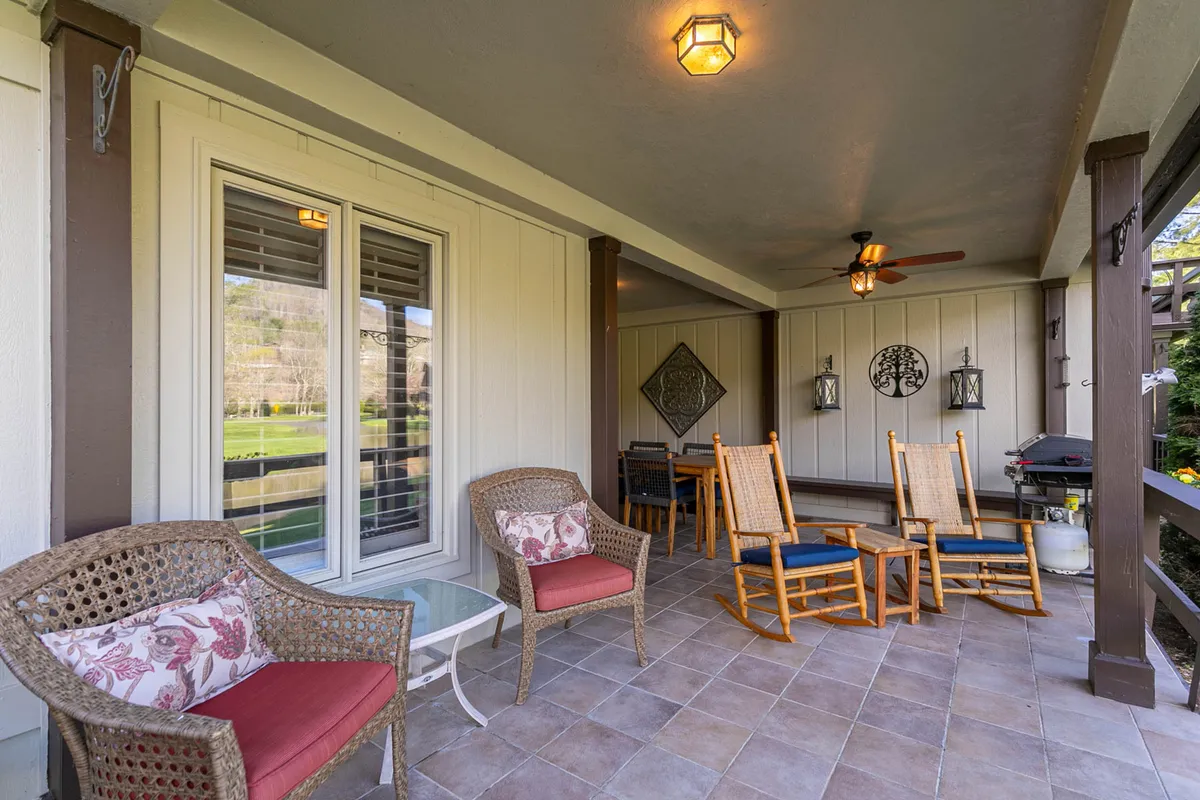 A cozy outdoor seating area features rocking chairs, woven chairs, and a coffee table set atop a covered porch. Decorative lanterns add a warm touch, and the arrangement invites guests to relax, socialize, and enjoy scenic views beyond the patio.