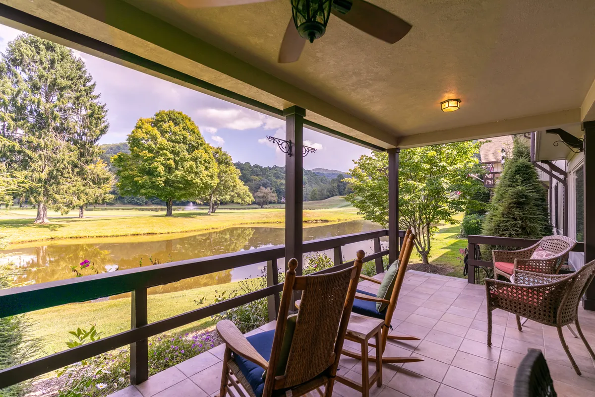 This exterior image features classic rocking chairs arranged on a porch, facing a tranquil pond surrounded by lush greenery and distant trees. The scene evokes a sense of peaceful relaxation, ideal for enjoying quiet mornings, reading, or simply taking in the natural surroundings.