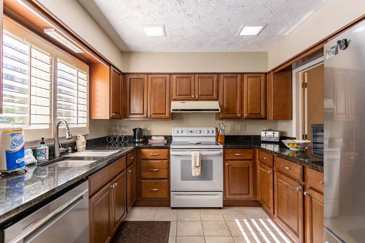 A bright and cheerful kitchen featuring granite countertops, wooden cabinets, and sleek stainless steel appliances. Sunlight streams in through a window with classic shutters, illuminating the spacious work surfaces and highlighting the kitchen’s modern yet welcoming design.